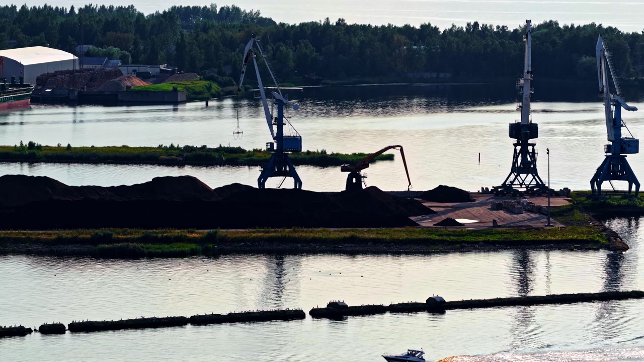 Harbor cranes stand beside calm water with cargo vessel and industrial shoreline in soft light
