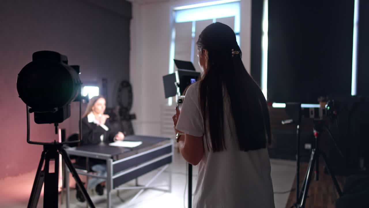 Long-haired brunette in white t-shirt starts to take a video of a woman sitting at desk. Bloggers create content for the blog.