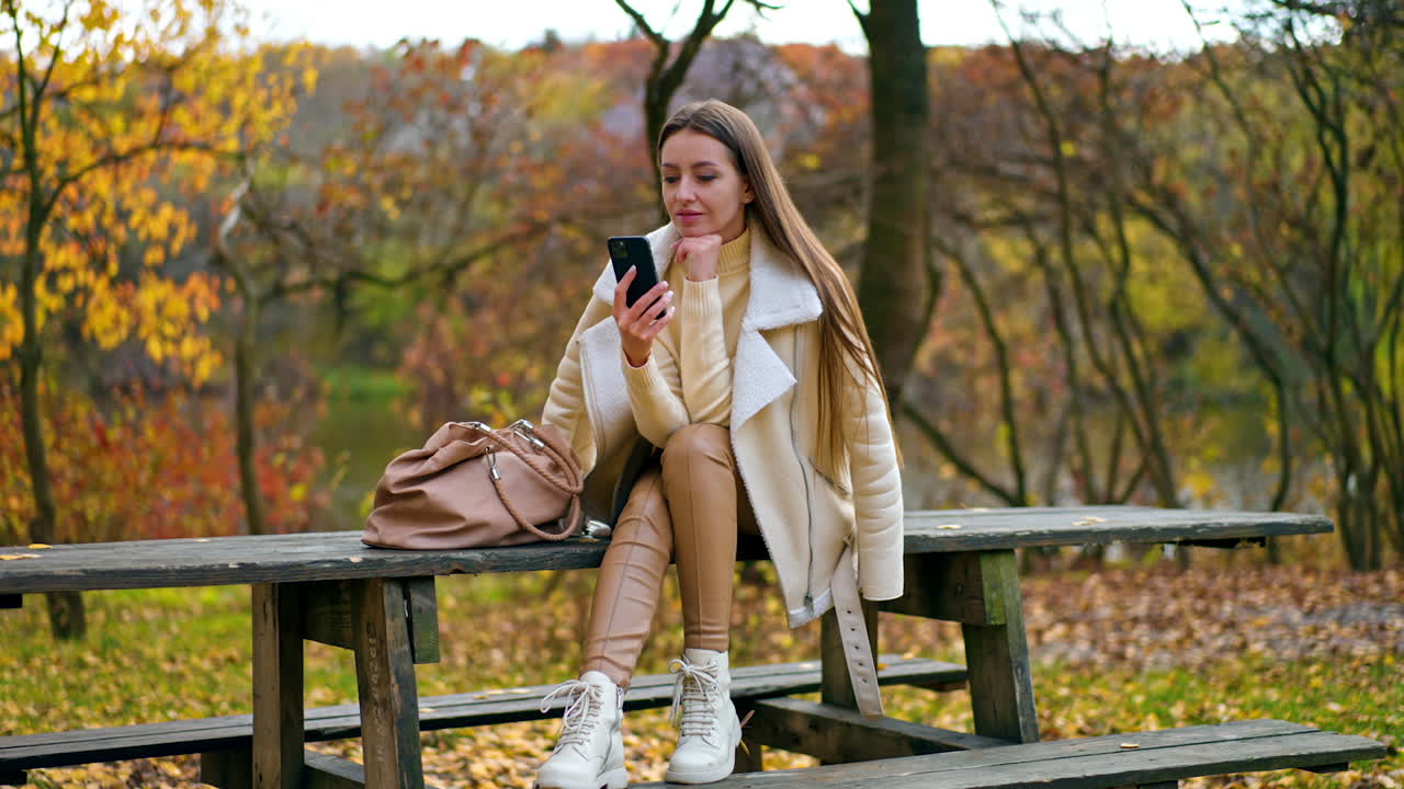 Pensive brunette sitting on the wooden table outdoors looking at her smartphone. Woman having rest in autumn park using gadget.