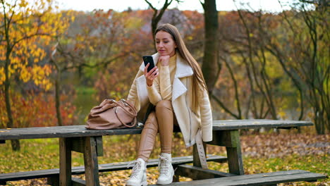pensive brunette sitting on the wooden table outdoors looking at her smartphone. woman having rest in autumn park using gadget.