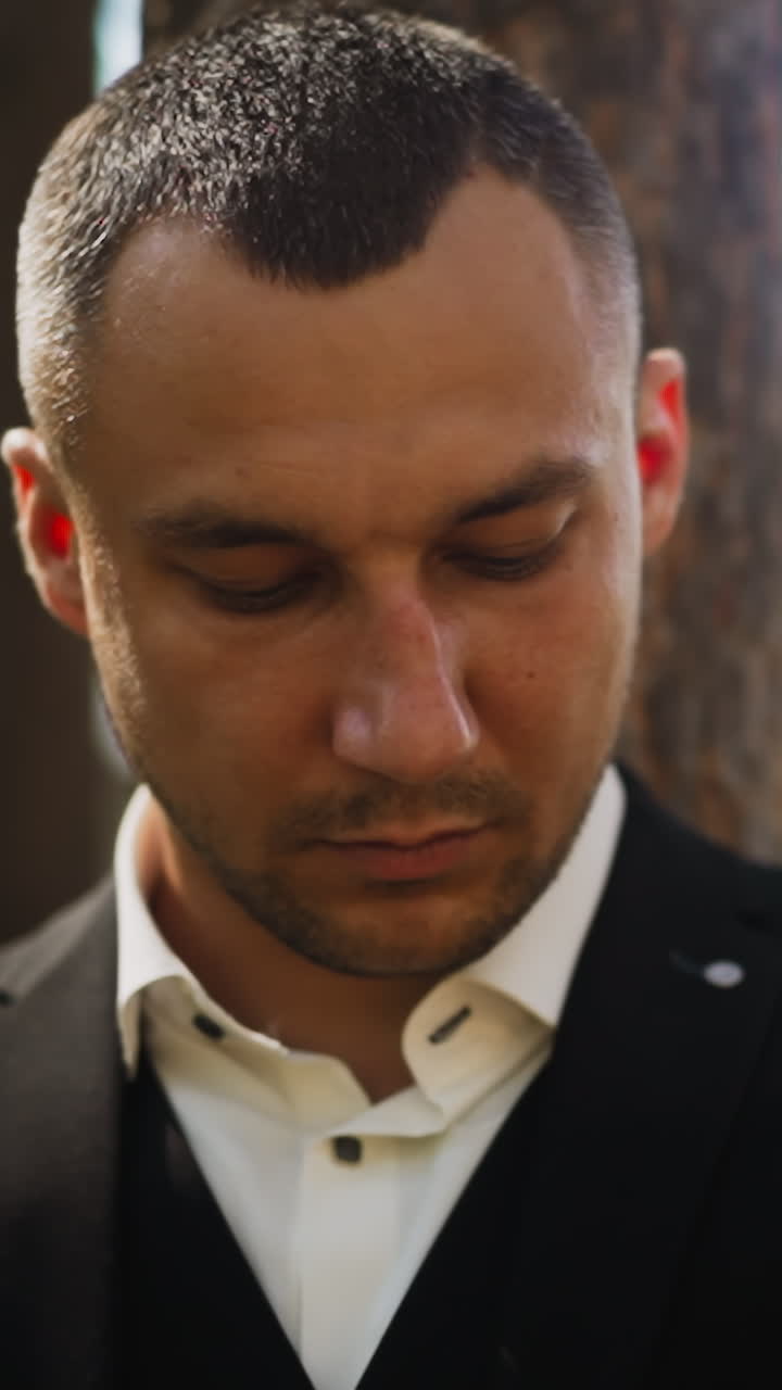 Concentrated gentleman with three day stubble wearing stylish black costume with white shirt looks downward standing in city park close view slow motion