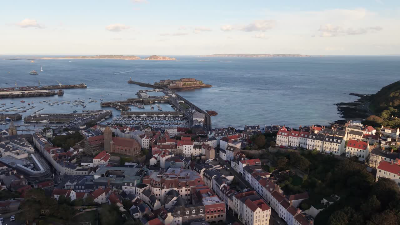 Reveal drone footage of the rooftops of St Peter Port Guernsey looking towards harbour and Castle Cornet Herm Sark and Jethou in the distance in bright afternoon sunshine on calm day
