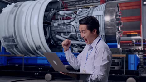 side view of an asian male worker with his laptop with airplane engine maintenance conducted, analytics checking the turbine, he raises his fist up with screaming goal after typed on the laptop