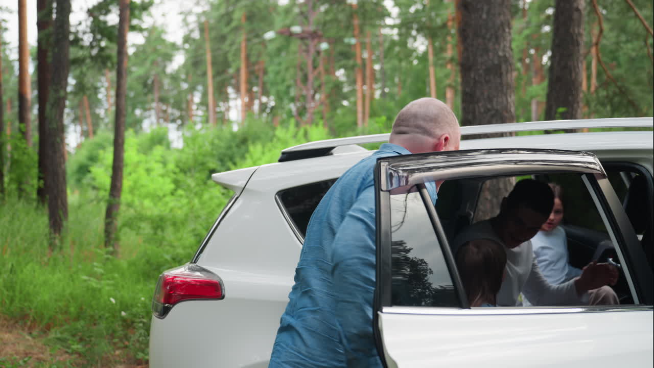 Father in blue shirt gently helps kids into white car parked among tall trees in forest, smiling as sunlight filters through greenery, calm family travel scene showing care, and summer atmosphere