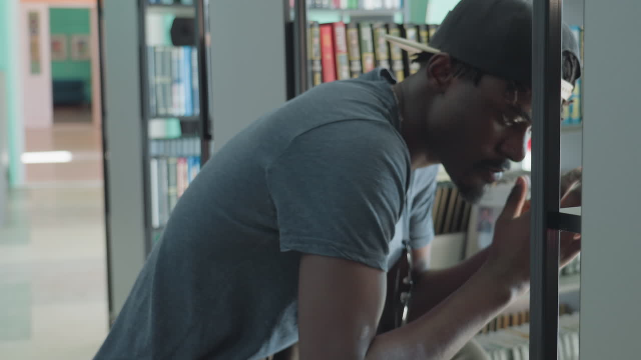 Giovane uomo con camicia grigia casual e berretto all'indietro accovacciato vicino allo scaffale di una biblioteca, che cerca ed esamina libri con espressione pensierosa, circondato da dorsi di libri colorati in un ambiente interno tranquillo