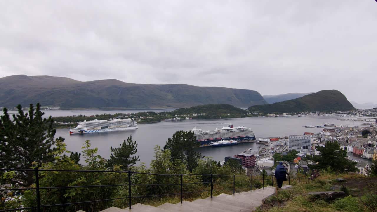 Person walking up a hill overlooking Ålesund harbor with cruise ships and scenic mountains