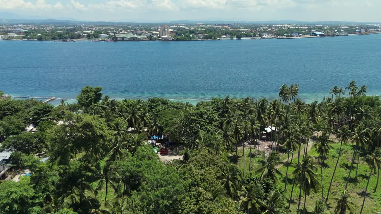 Aerial drone footage flying over a tropical resort and residential area with modern roofs and lush palm trees. Across the deep blue bay, a busy coastal city and industrial port fill the background