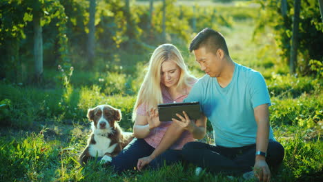 A-Young-Couple-Is-Sitting-Near-A-Vineyard-Using-A-Tablet-Next-To-Them-Is-Their-Dog-Private-Tasting-A