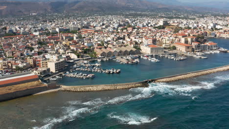 waves splashing at the port of chania in crete island, greece with view of marina and seafront houses