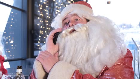 Santa Claus is Talking on a Smartphone he Sitting in an Irish Pub and Relaxing Santa Claus is Talking on a Smartphone he is Sitting in an Irish Pub and Relaxing Young Santa Claus Uses Gadget Concept of Celebration and New Year Gifts
