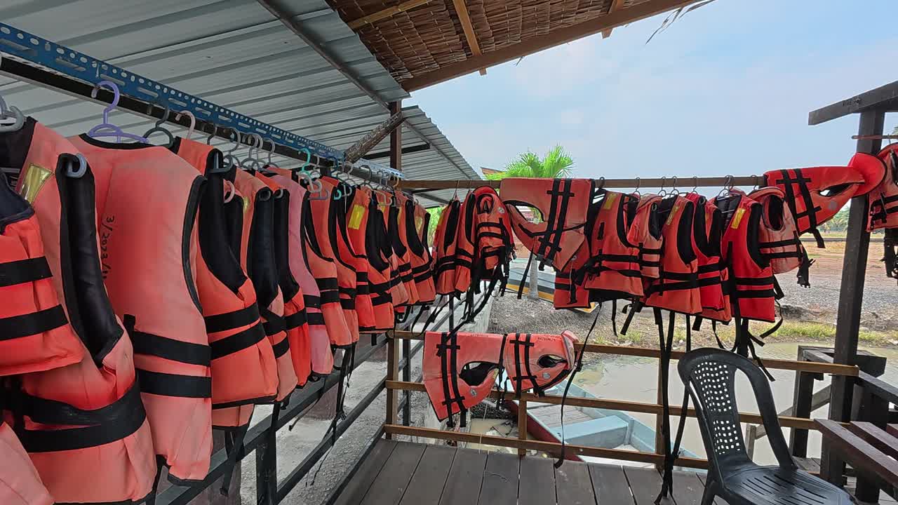 Un assortiment de gilets de sauvetage orange usagés est suspendu et disposé sur une jetée en bois traditionnelle. Ces gilets sont des équipements de sécurité pour les habitants qui utilisent de petites embarcations pour leurs déplacements et activités quotidiens.