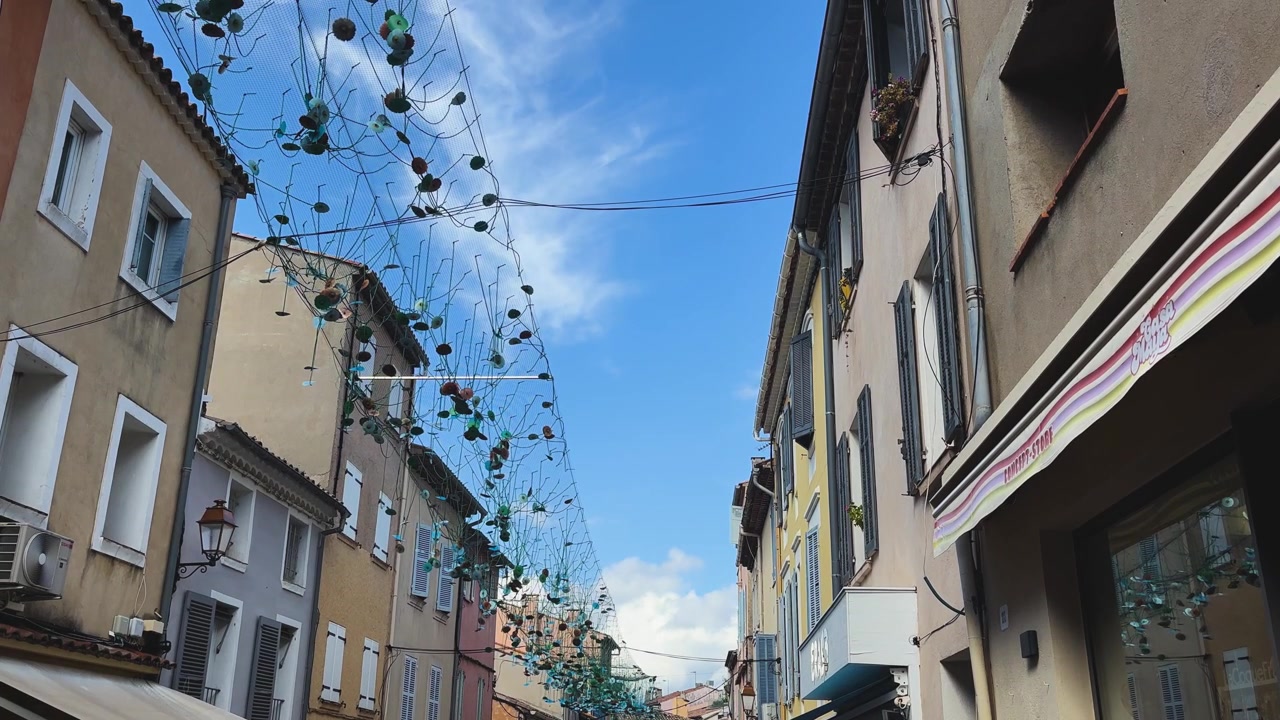Ornamenti colorati sospesi formano un baldacchino artistico sopra una strada stretta fiancheggiata da edifici color pastello sotto un cielo azzurro brillante