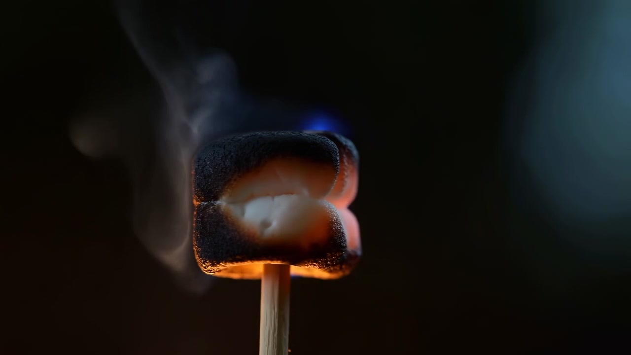Close-up van marshmallow op houten stok die brandt in donkere studio, met blauwe vlam, oranje gloed, opstijgende rook, smeltend oppervlak en verkoolde korst tegen diepzwarte achtergrond, met marshmallow, vlam.