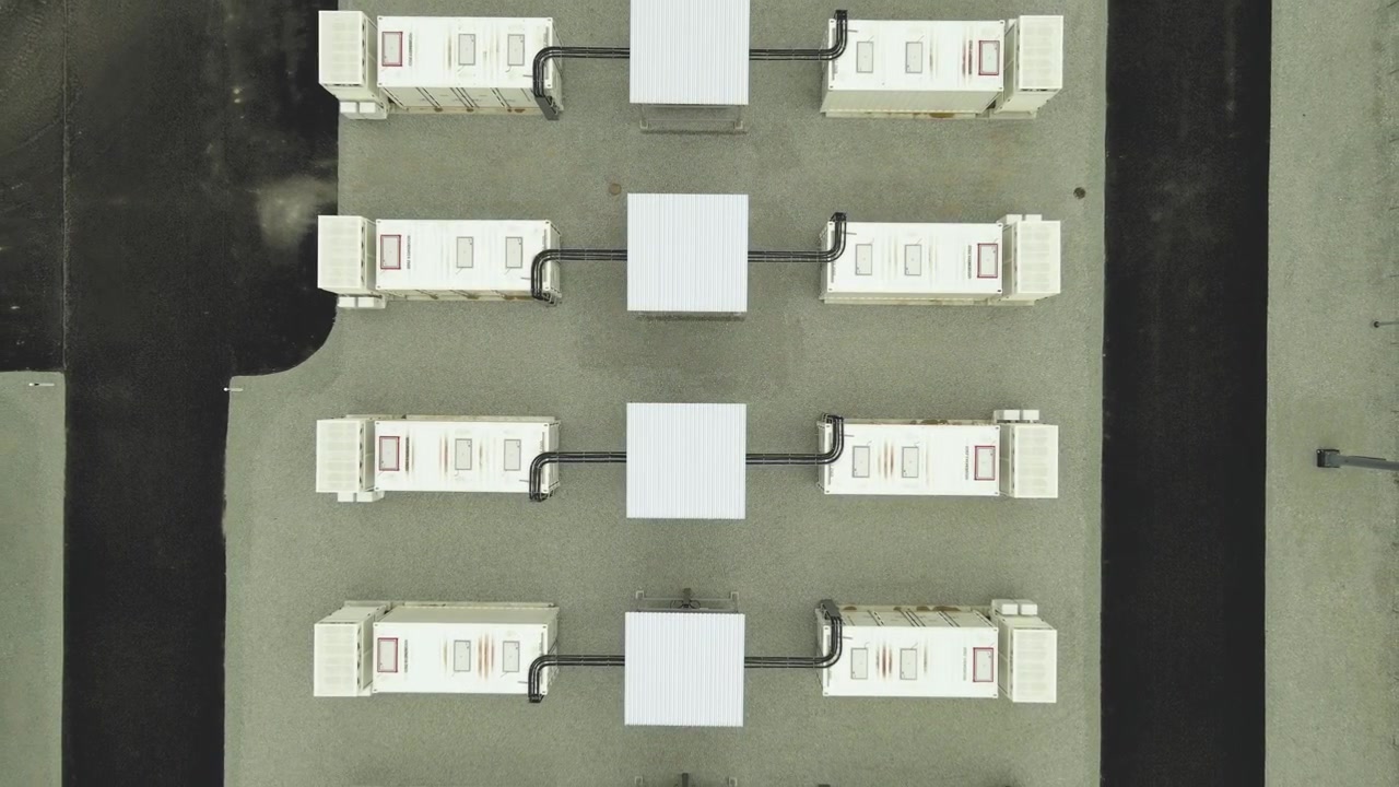 Vista aerea dall'alto che sorvola un moderno impianto BESS con grandi contenitori che immagazzinano energia rinnovabile per la rete elettrica e l'industria.