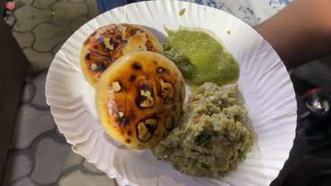 man holding a plate of savoury litti chokha served with baingan bharta or roasted mashed eggplant and green spicy chutney on a paper plate