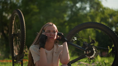 young lady seated next to her upside-down bike, looking at her phone with a tired and frustrated expression, surrounded by lush greenery and trees, capturing a moment of contemplation