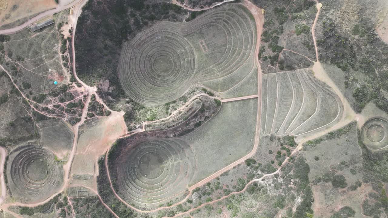 Aerial drone view of Moray circular agricultural terraces in Peru, sunlit stone structures beneath lightly scattered clouds