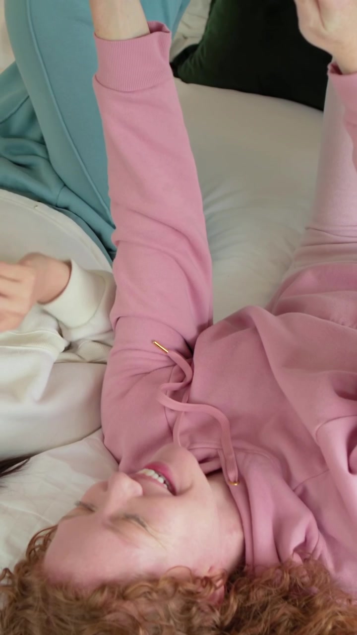 Vertikal video: Leende kvinner i rosa hettegenser og turkise bukser holder nettbrett etter å ha sett på i sengen