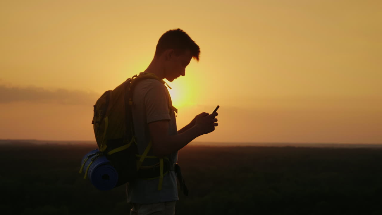 Un giovane turista con uno zaino usa lo smartphone in cima a una montagna, tra i raggi del tramonto.