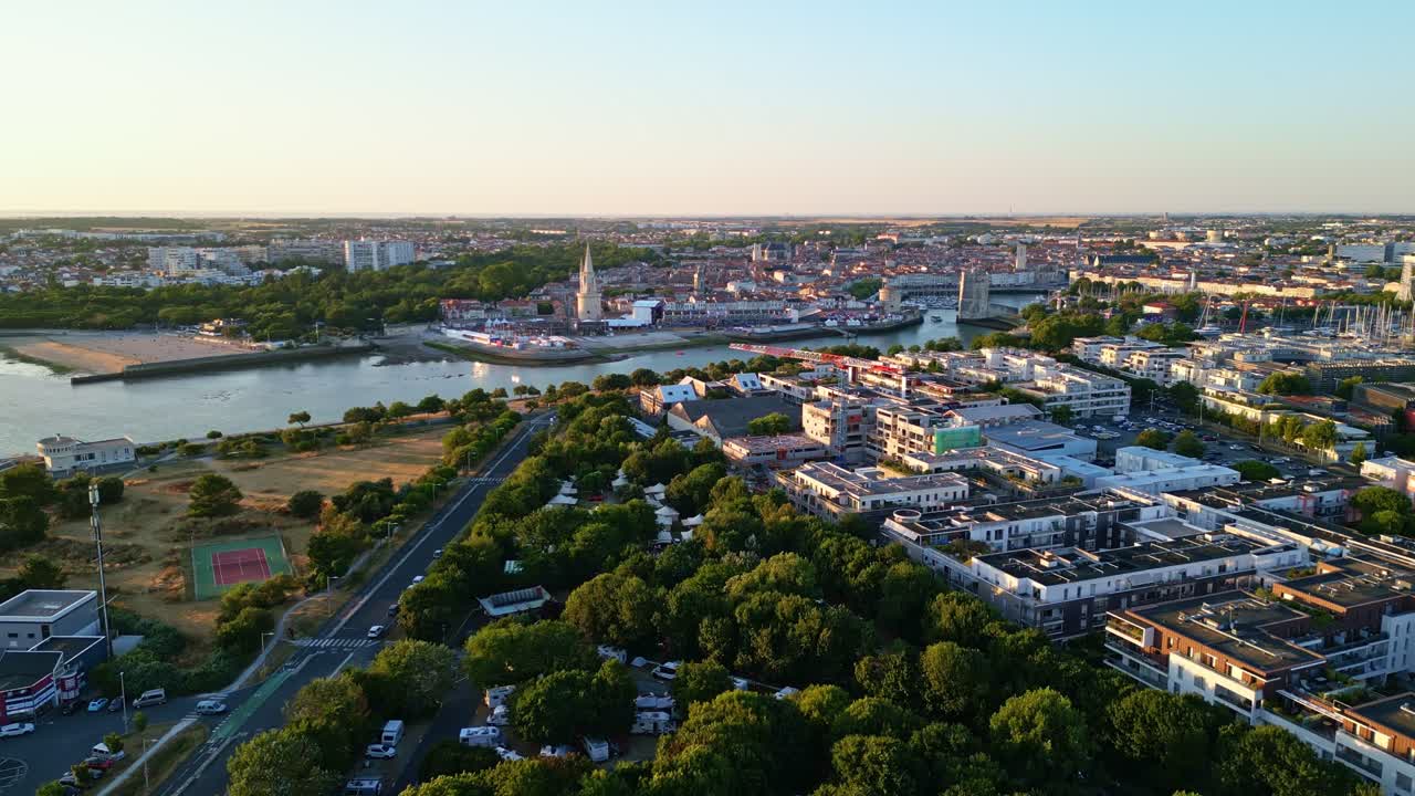 Drone shot over La Rochelle at sunset, showing a tree-lined avenue, tennis court, Lantern Tower, port area with festival tents, and cityscape in the distance - France