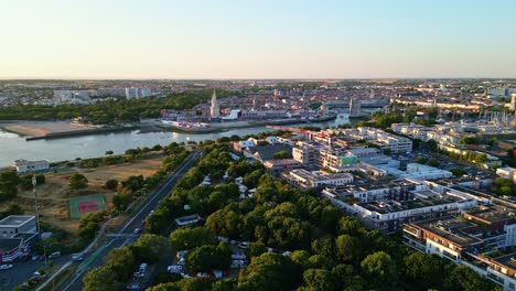 drone shot over la rochelle at sunset, showing a tree-lined avenue, tennis court, lantern tower, port area with festival tents, and cityscape in the distance - france