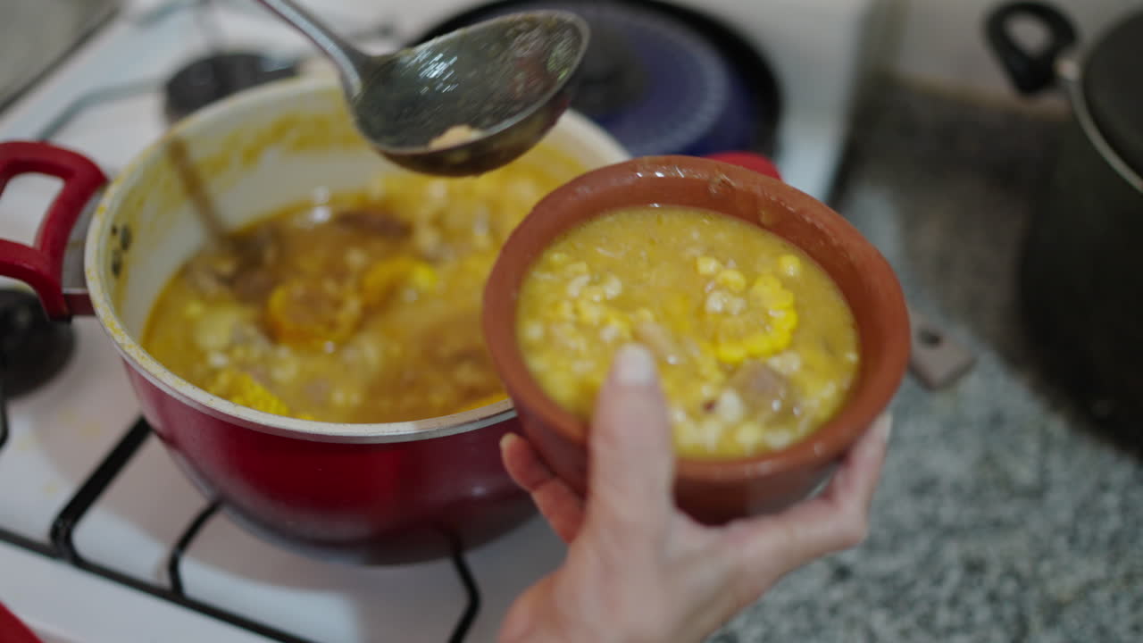 Hand serving thick Locro Argentino corn stew from pot into traditional clay bowl.