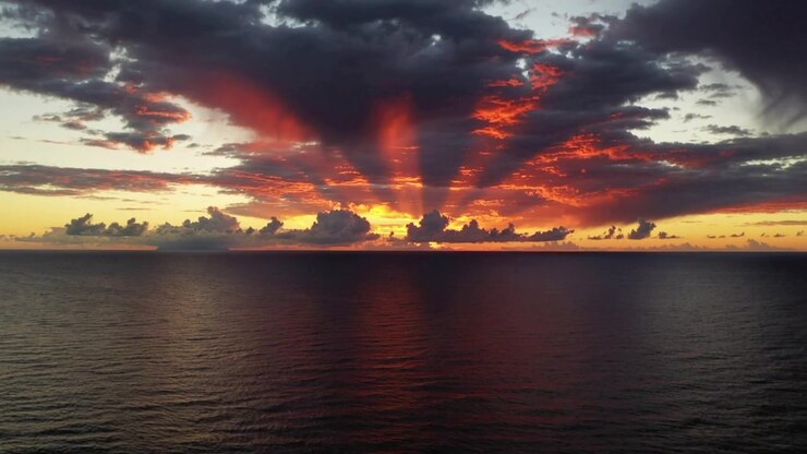 Sol naciente detrás de un denso banco de nubes en el horizonte, proyectando rayos crepusculares y reflejando una franja cálida en el océano.