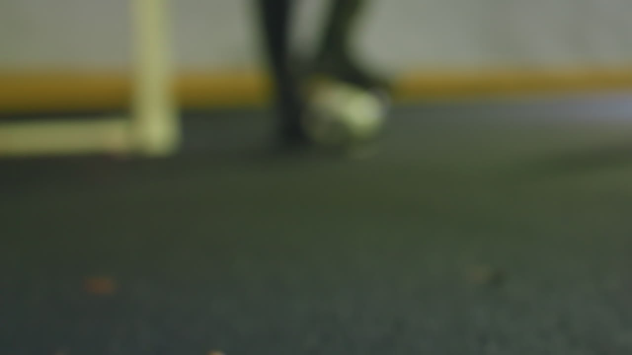 primer plano de guantes de portero acostados cerca del poste de la portería en césped artificial rodeado de hojas secas esparcidas, el fondo borroso muestra a un jugador de fútbol equilibrando la pelota bajo los pies por la noche