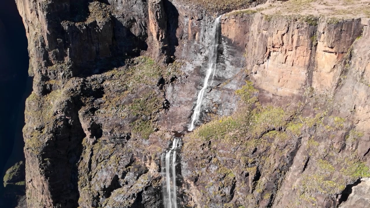 Una cascata alta e stretta precipita da una ripida scogliera rocciosa accanto all'apertura di un canyon scuro, mostrando aspre texture di pietra, vegetazione rada e una drammatica luce naturale del giorno.