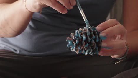 close up on caucasian woman's hands, painting and decorating a pine cone with blue light colour, udsing a brush 120fps