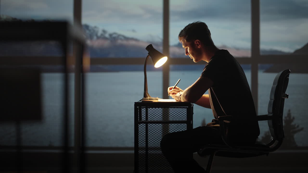 Serious concentrated young male student studying at home office sitting in cozy workplace at night under lamplight. A young man thinks creates a creative project and writes it down with a pen.