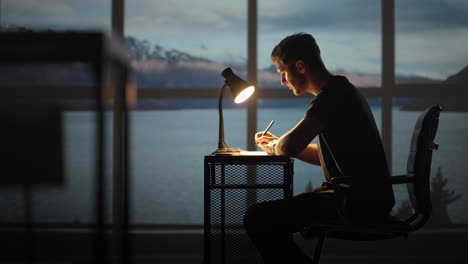 serious concentrated young male student studying at home office sitting in cozy workplace at night under lamplight. a young man thinks creates a creative project and writes it down with a pen.