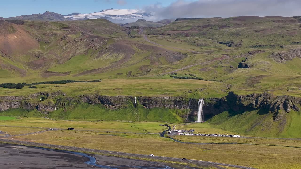 Aerial panning shot of seljalandsfoss iceland during sunlight with green facade and snowy peaks. Wide shot