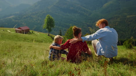 Relaxed-family-sitting-grass-mountain-slope-at-summer.-Parents-hugging-children.