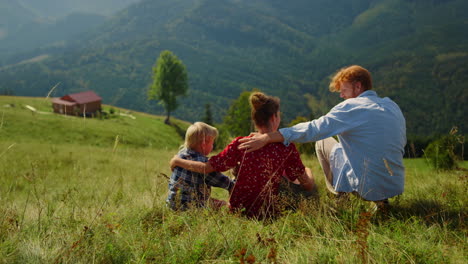 relaxed family sitting grass mountain slope at summer. parents hugging children.
