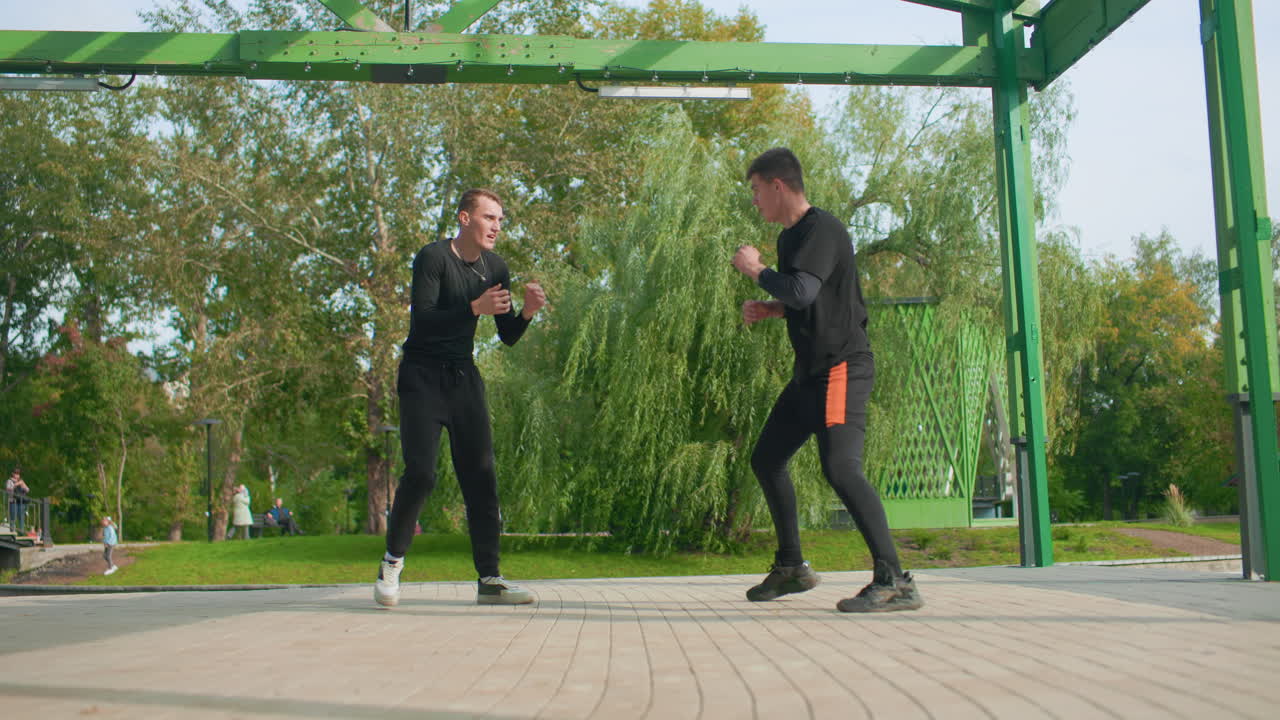 To mænd øver nærforsvar under en metalpergola i en bypark, hvor de udfører clinch- og kontramanøvrer, grebskontrol og momentændringer med et taktisk fokus, realistisk scenarietræning og intensitet.