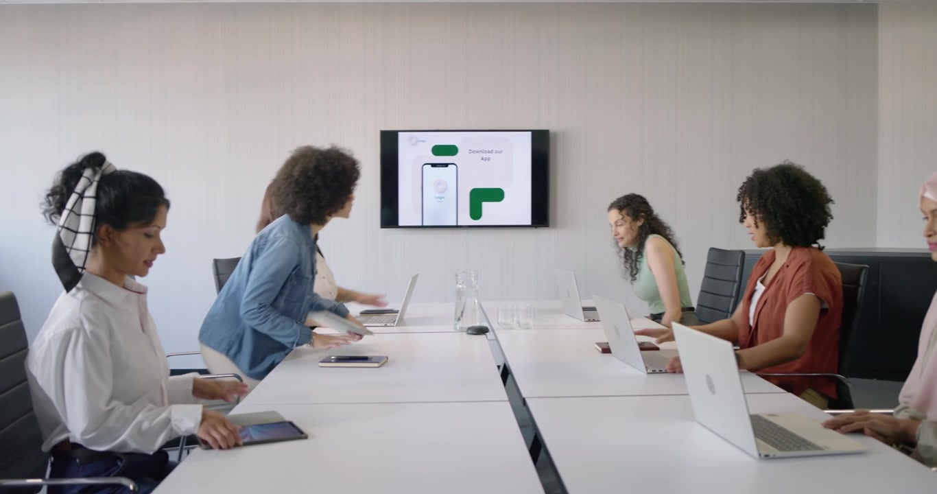 Diverse female coworkers entering meeting room placing laptops on table by screen preparing meeting