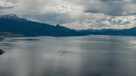 thick, white-gray clouds hanging above the dark waters of the hardanger fjord, norway. rays of sun are piercing through the clouds, ripples running on the water surface. mountains tower on the horizon