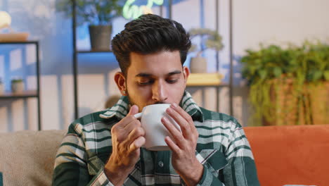 Handsome-smiling-indian-man-drinking-a-cup-of-coffee-or-herbal-tea-sitting-at-home-couch,-break-time