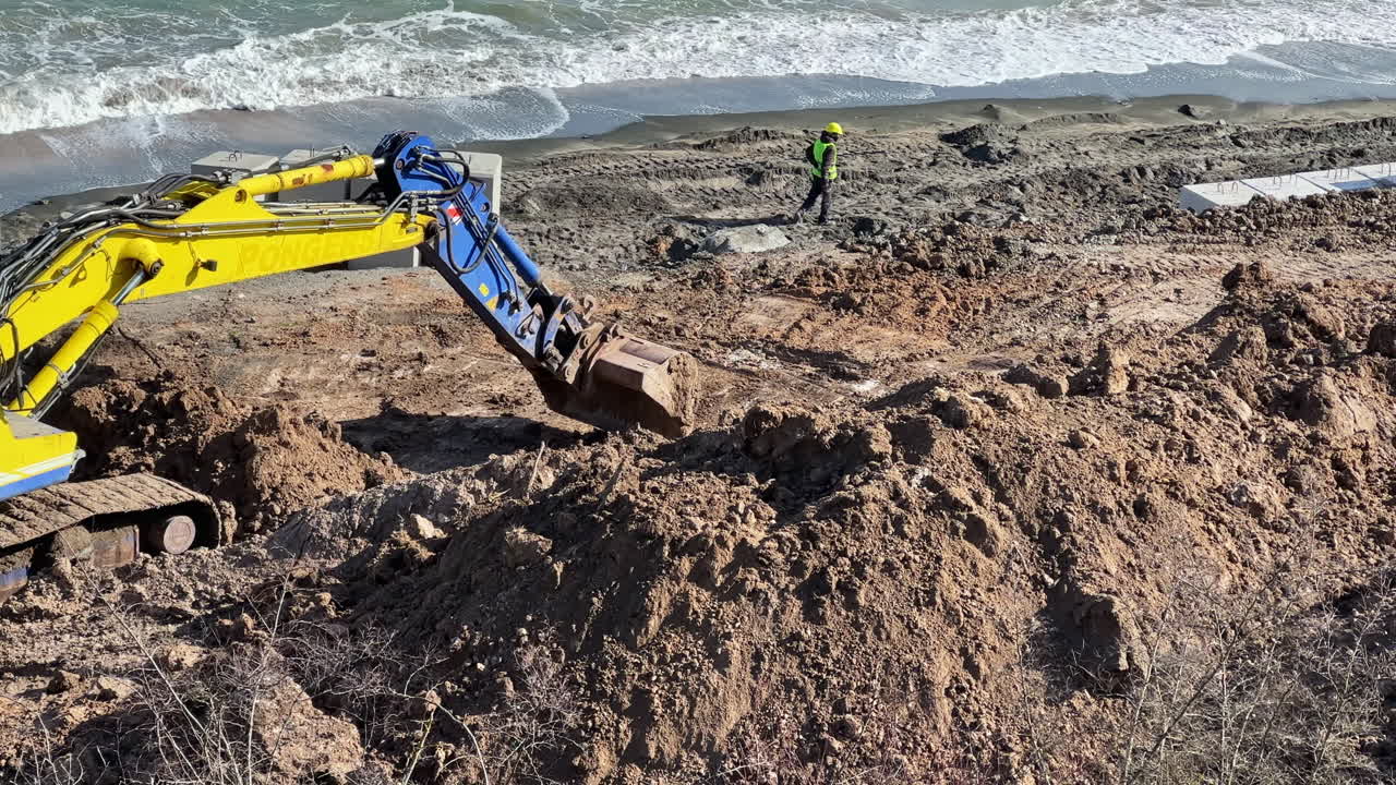 Lavori di costruzione si svolgono in un cantiere costiero mentre i macchinari spostano terra vicino alla battigia in una giornata di sole.