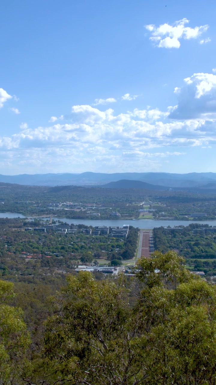Uma panorâmica ampla da cidade de Canberra, do Lago Burley Griffin e de cadeias de montanhas distantes.