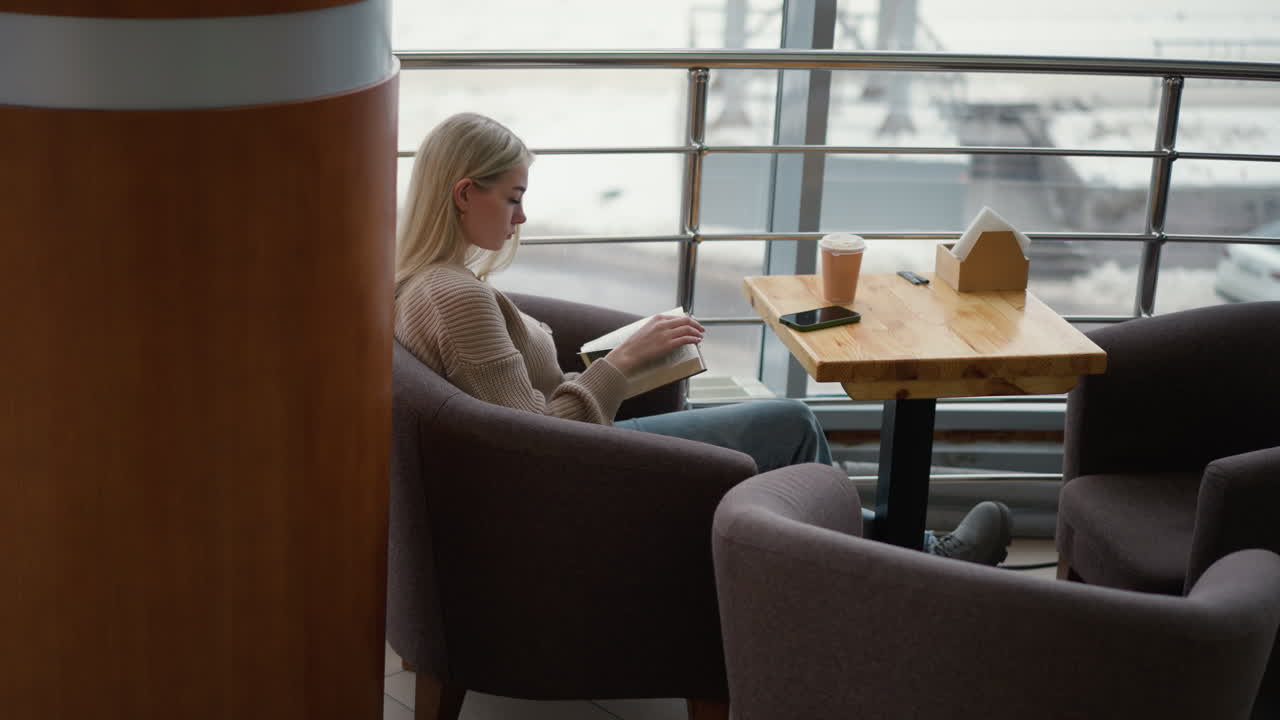 vista lateral de una mujer sentada en un acogedor café, leyendo un libro pensativamente con una taza de café y un teléfono en la mesa, relajada y centrada en la lectura mientras se ve la vista urbana afuera