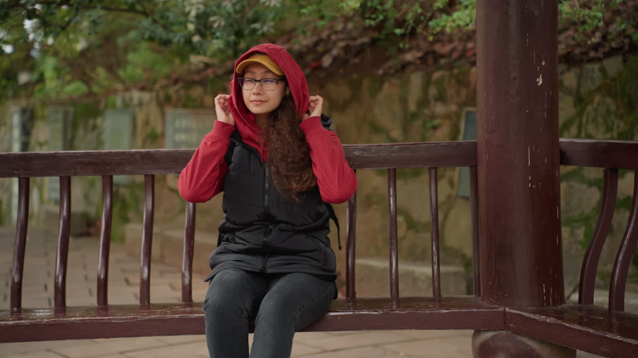 Mujer joven con sudadera roja sentada en un banco de madera de un cenador, mirada pensativa a través de las gafas, chaleco acogedor sobre la chaqueta, fondo de follaje otoñal, luz natural suave, ambiente tranquilo y reflexivo, vida lenta.