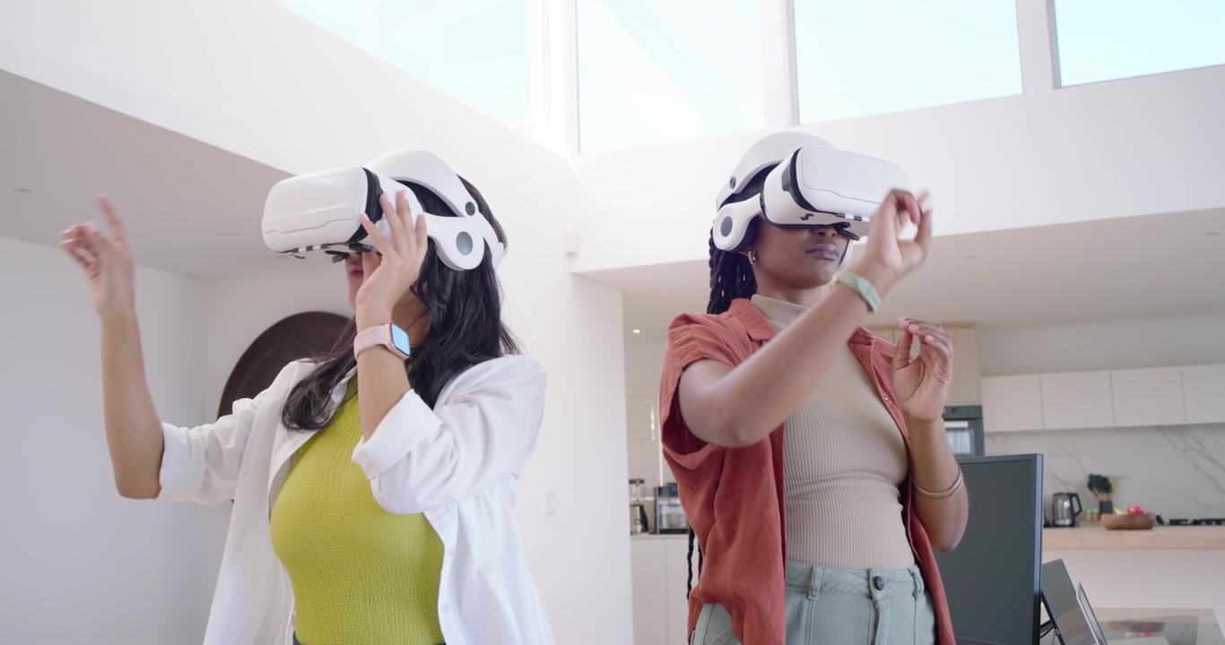 Due donne diverse che sistemano i visori VR, gesticolando per una dimostrazione in cucina mentre una collega usa un laptop.