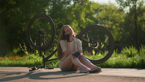 young lady seated near her upside-down bike, answering a phone call with a concerned expression, surrounded by lush greenery and trees in an outdoor setting