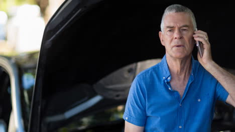 senior caucasian man using smartphone beside open bonnet of broken-down car