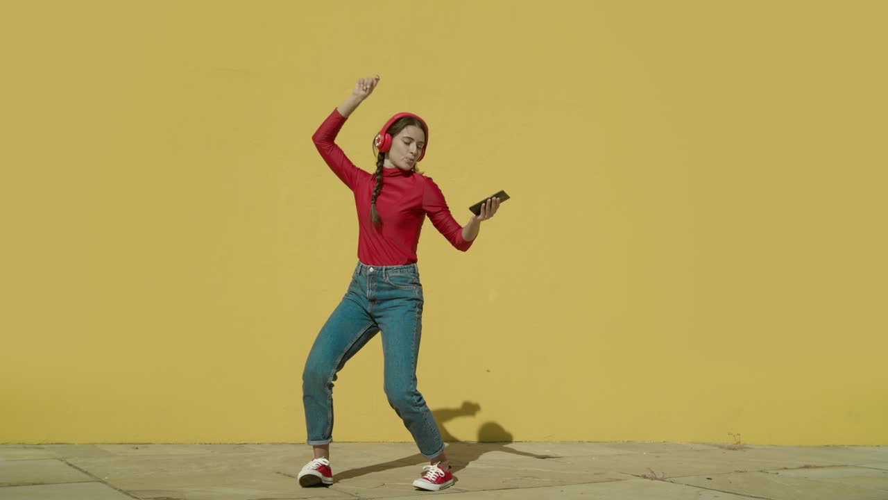 Young Happy Latin Woman makes dancing cool moves wearing a red shirt and jeans and red converse shoes with a yellow background wall on a sunny day wide-angle shot in slow motion