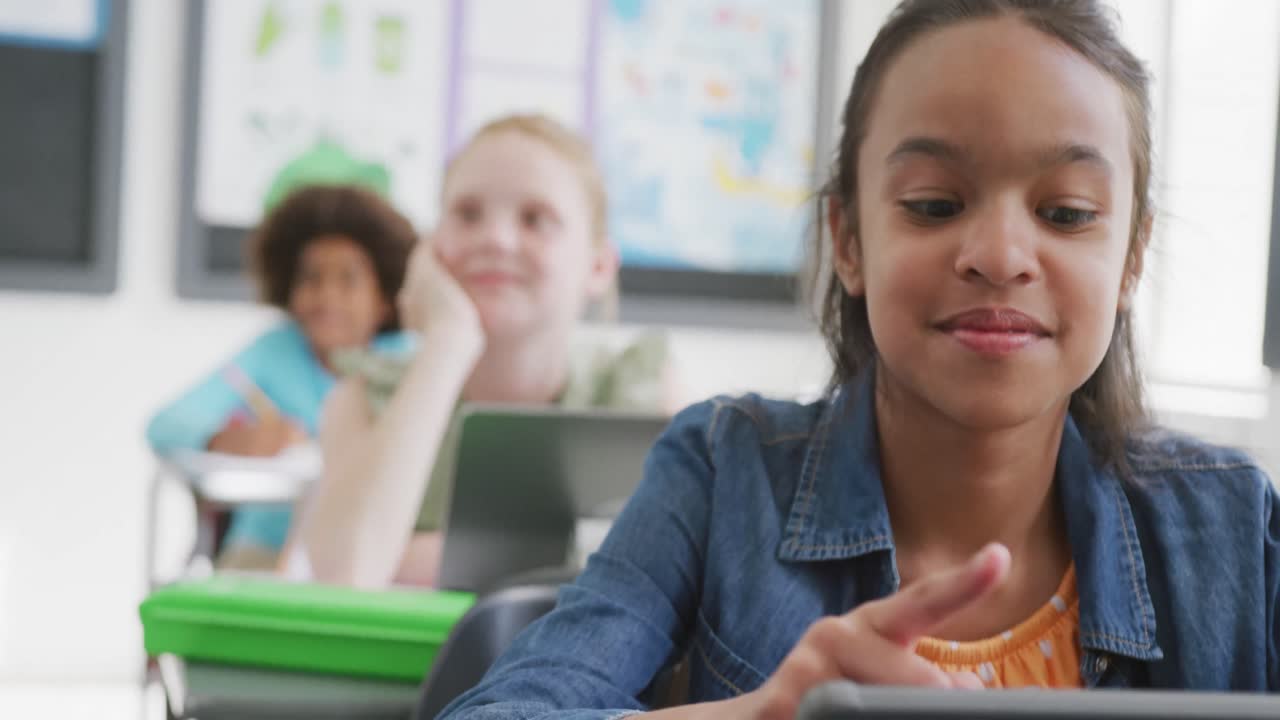 Video of smiling biracial schoolgirl sitting at desk using tablet in class, copy space
