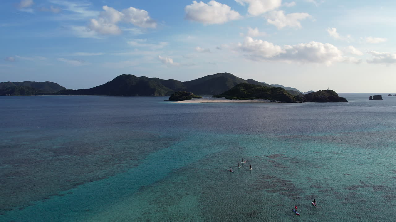 A drone descends from 80m to 3m over Zamami, revealing aquamarine waters, SUP paddlers, and pristine white sand beaches in this tropical island paradise