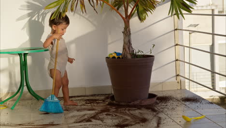 little latin baby boy wearing a baby romper cleaning the floor with a blue toy broom after soiling it with dirt from a plant pot